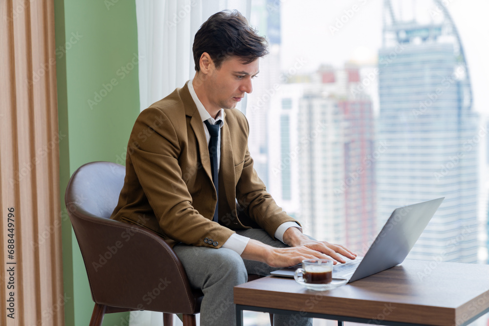 Portrait of skilled businessman working with laptop near window with skyscraper. Professional executive manager typing searching analyzing data on table with laptop and coffee cup. Ornamented.