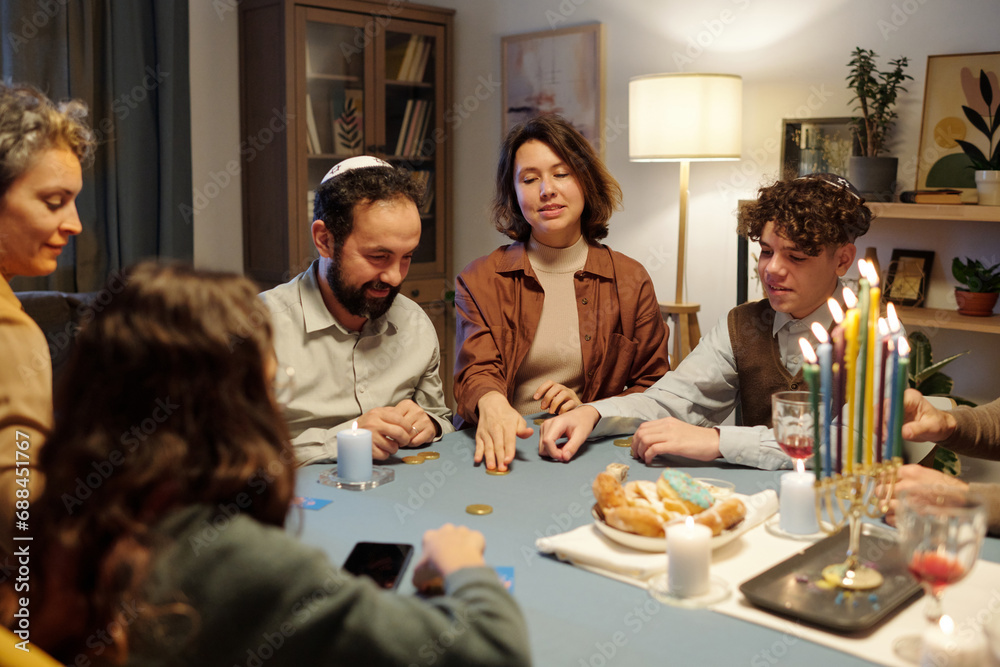 Young woman sliding coin on table while playing leisure game with members of her family sitting by table with menorah candlestick Young woman sliding coin on table while playing leisure game with members of her family sitting by table with menorah candlestick
