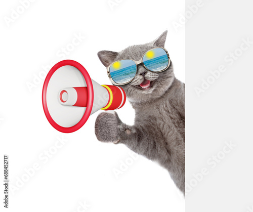 Happy cat wearing mirrored sunglasses looks from behind empty board and talks into megaphone. isolated on white background