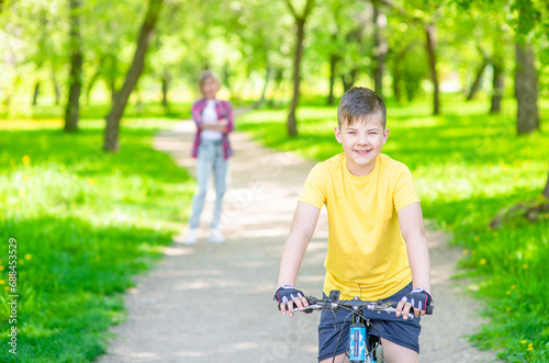 Happy young boy rides on a bicycle in summer sunny park. Proud mom stands behind on the background