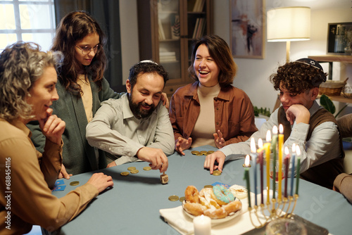 Young cheerful woman laughing while sitting next to mature man spinning small dreidel during leisure game among family members