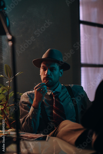 Young pensive retro man in hat and formalwear holding cigarette and thinking of something while sitting in dark office or room