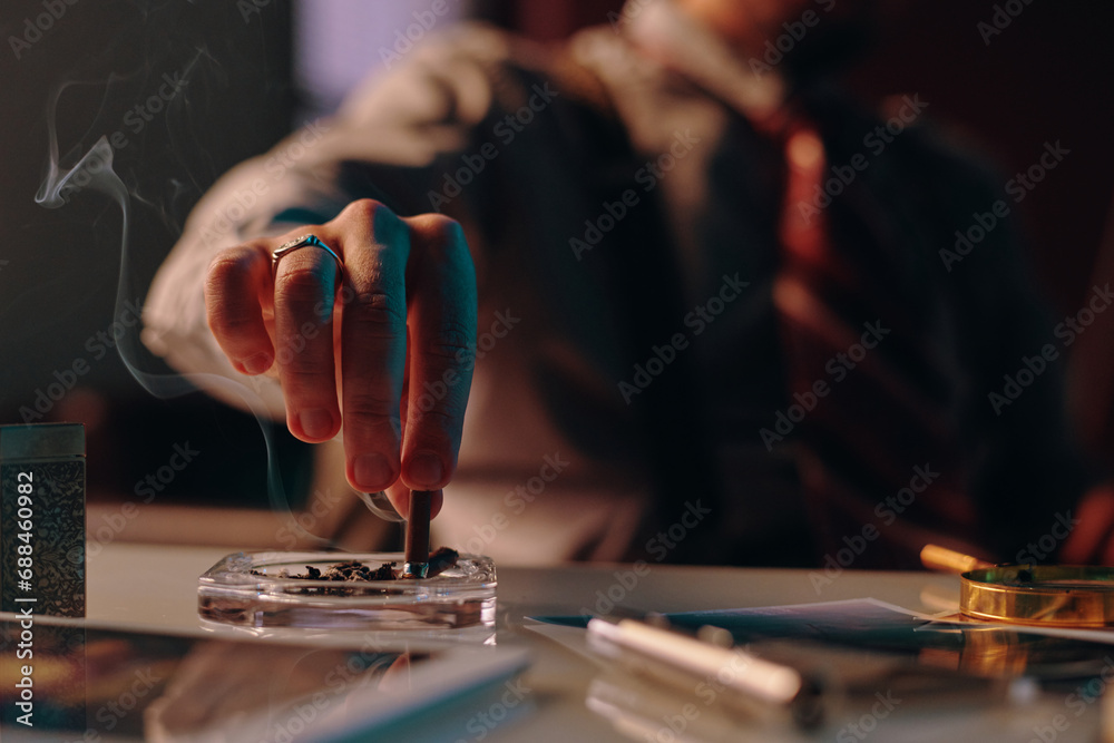 Close-up of hand of young man stubbing butt of cigarette in ashtray standing on table among business and office supplies Close-up of hand of young man stubbing butt of cigarette in ashtray standing on table among business and office supplies
