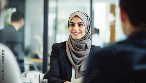 A young Muslim woman in a hijab sits at a table in an office, communicating with an interlocutor. photo