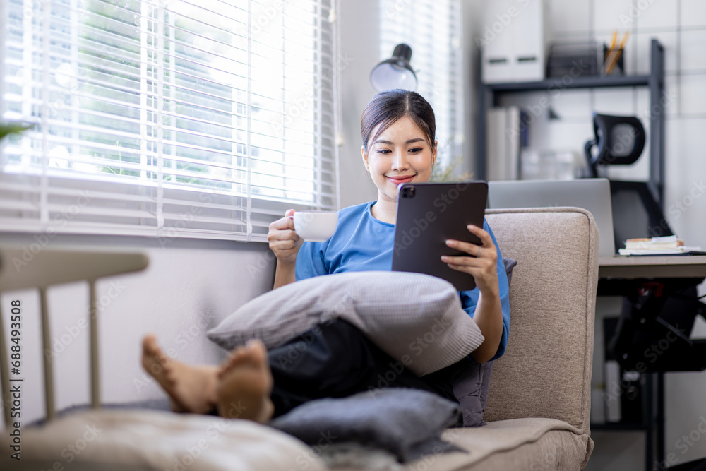 Asian young woman sitting on the yellow sofa using digital tablet and coffee cup and learning shopping or working online near window Asian young woman sitting on the yellow sofa using digital tablet and coffee cup and learning shopping or working online near window