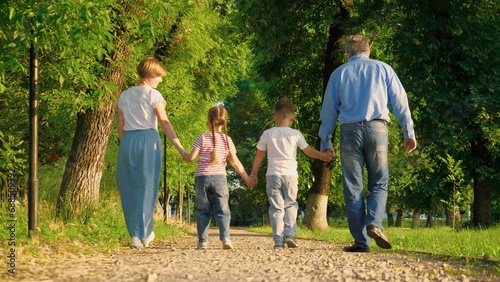 Small children walk with joint hands with pleased parents on park ground road