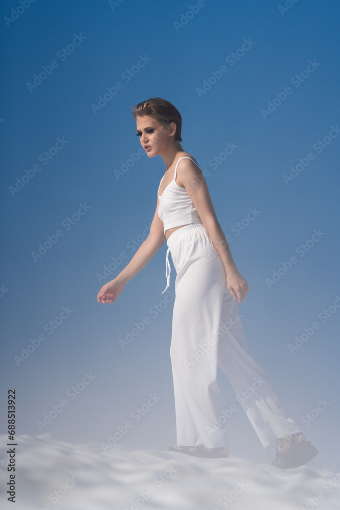 Beautiful blond woman dressed in white crop top, white pants and sandals. European female tourist walking through snow with light fog on sunny day with blue sky. Full length, side view Beautiful blond woman dressed in white crop top, white pants and sandals. European female tourist walking through snow with light fog on sunny day with blue sky. Full length, side view
