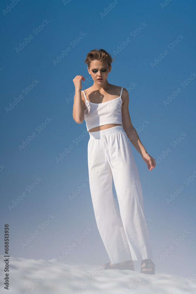 Woman walks through snow with light fog on sunny weather with blue sky. Romantic blond adult female looking down. Tourist wearing in white crop top, white pants and sandals. Full length, front view Woman walks through snow with light fog on sunny weather with blue sky. Romantic blond adult female looking down. Tourist wearing in white crop top, white pants and sandals. Full length, front view
