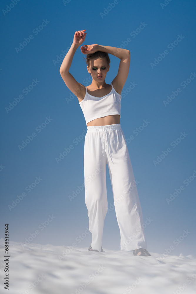 Obraz premium Sexual blond adult woman dressed in white crop top, white pants and sandals. European female tourist looking down, hands up, walking through snowfield with light fog on sunny day with blue sky