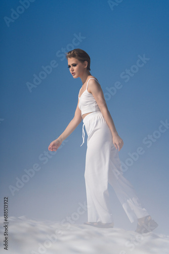 Beautiful blond woman dressed in white crop top, white pants and sandals. European female tourist walking through snow with light fog on sunny day with blue sky. Full length, side view