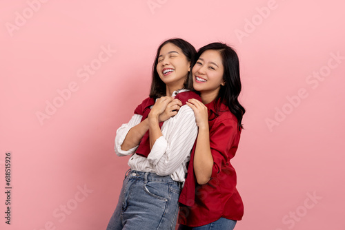 Two happy smiling Asian female friends hugging each other, wearing red and white shirts, standing isolated on pink background.