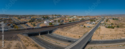 Aerial view of a curving highway through a hillside cityscape with beige buildings, green spaces, and distant mountains under a clear blue sky.
