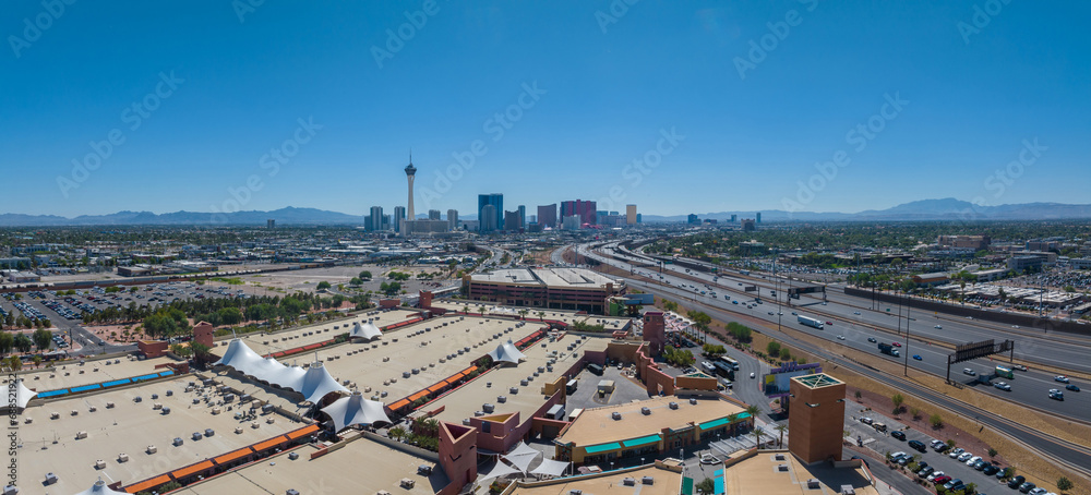 Aerial view of Las Vegas cityscape featuring the iconic Stratosphere Tower, diverse architecture, bustling roads, and green spaces against a mountainous backdrop in clear daylight.