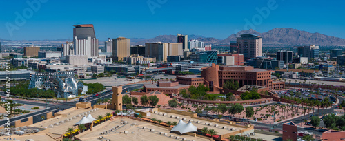 Aerial view of Las Vegas with modern high-rises, unique architecture, green parks, and a circular complex, set against a mountainous backdrop under a clear blue sky.