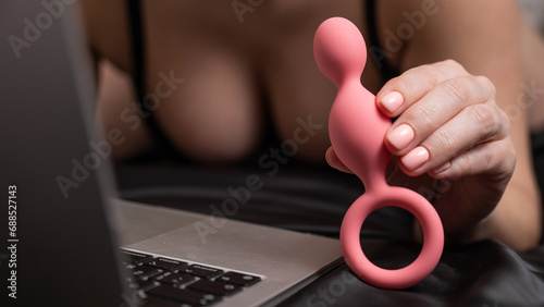 Woman holding pink anal beads next to laptop while lying on black sheet.  photo