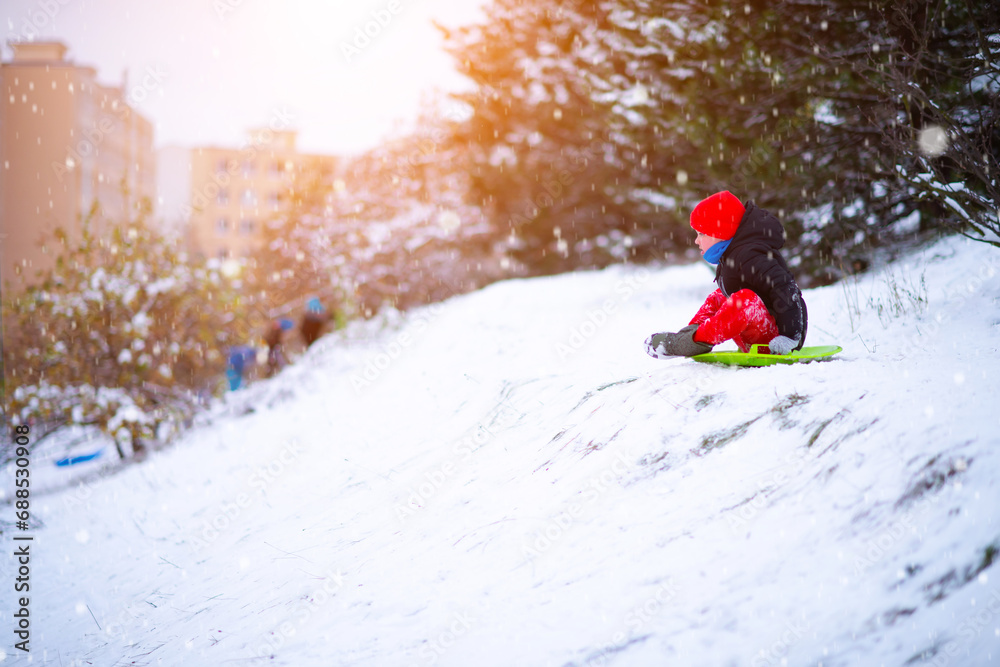 A little boy in a red hat and red pants slides down a snow slide on a round snow board