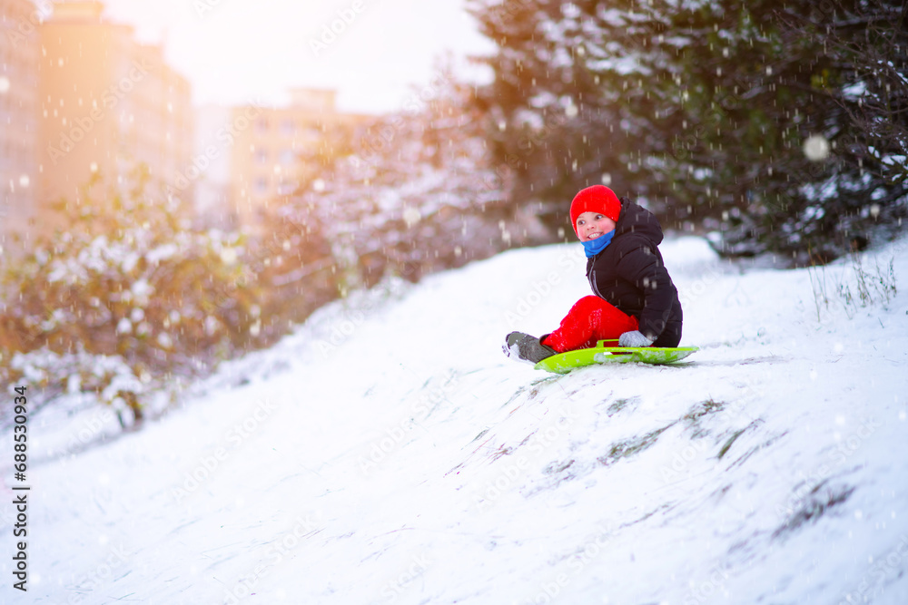 A little boy in a red hat and red pants slides down a snow slide on a round snow board