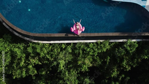 Top view of millennial young woman swim with inflatable pink flamingo in infinity pool in tropical jungle. Female travel blogger sunbath relax rest in luxury villa on summer vacation trip, pool party  photo
