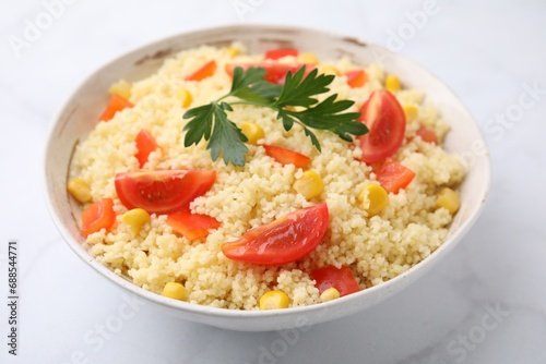 Tasty couscous with parsley, corn and tomatoes in bowl on white marble table, closeup