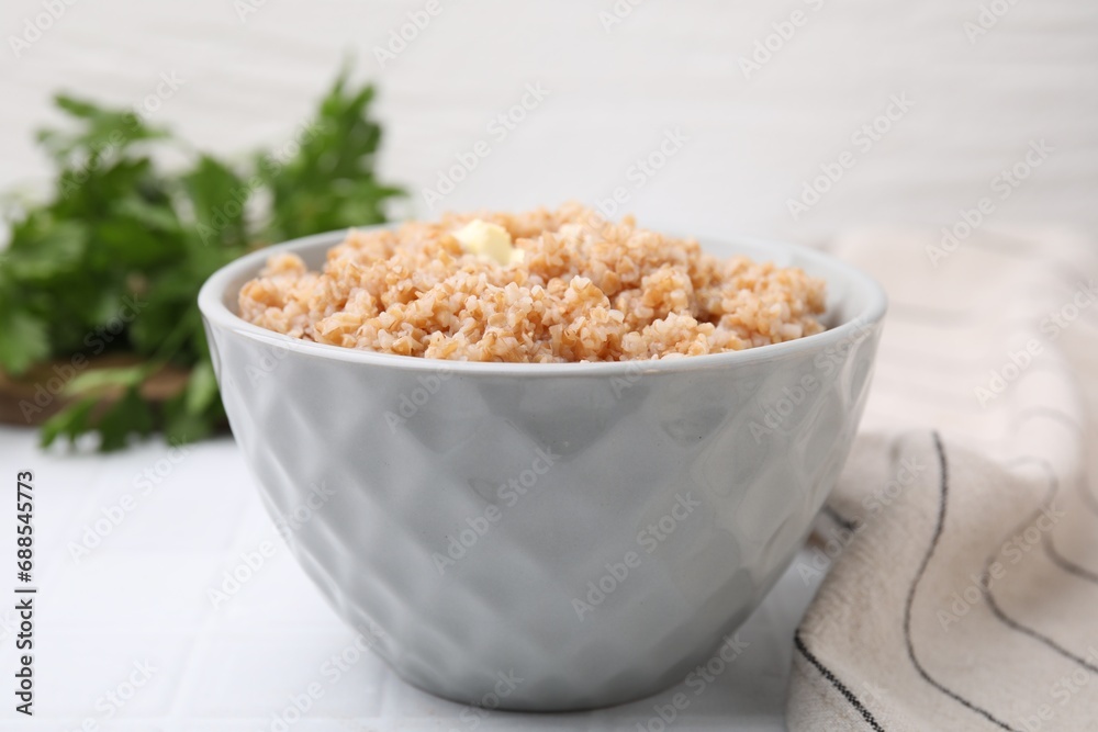Tasty wheat porridge with butter in bowl on white tiled table, closeup