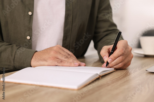 Young man writing in notebook at wooden table, closeup