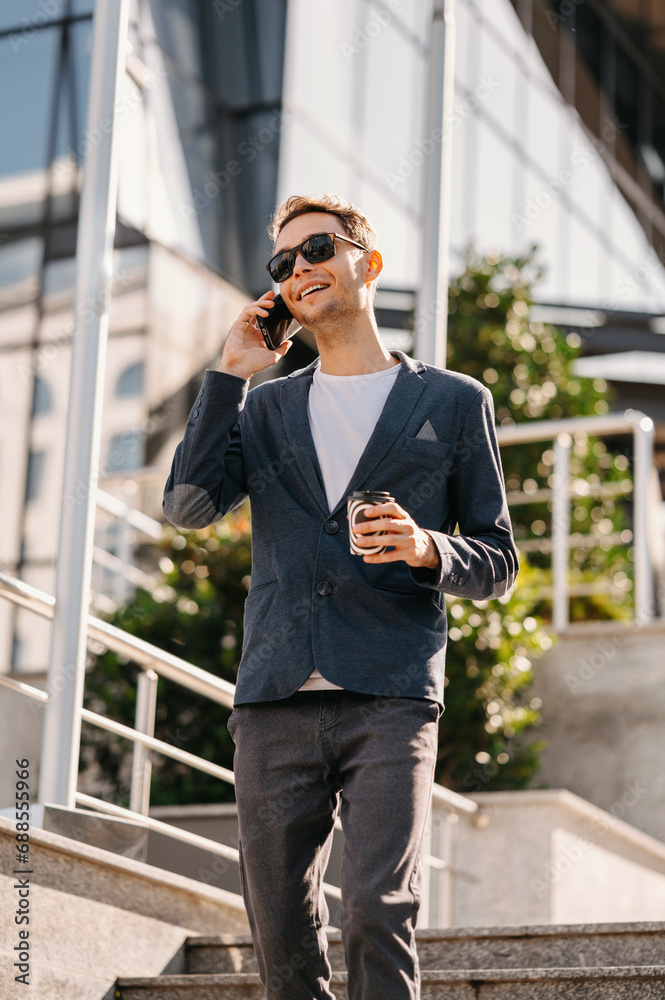 Walking cheerful office worker talking on smartphone outdoors and drink cup of coffee to go.