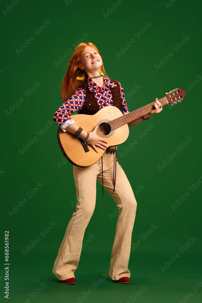 Full-length portrait of beautiful young woman, teenager looks like cowgirl and paying guitar against green studio background.