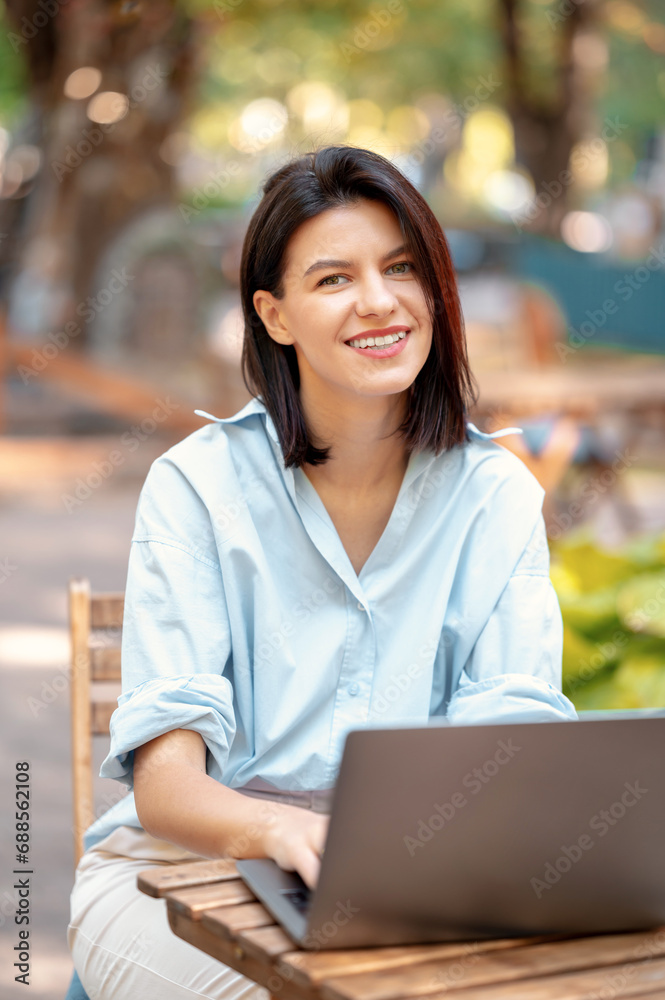 Vertical image of charming young woman in casual sitting outdoors at cafe and working at laptop remote. Vertical image of charming young woman in casual sitting outdoors at cafe and working at laptop remote.