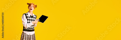 Banner. Adorable, pleasantly shocked young girl, student pointing on tablet with blank screen against yellow background with negative space for text. photo