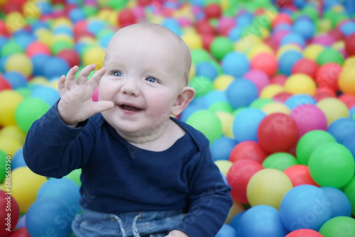 A cute eared baby among the colorful balls smiles and waves his hand. Cute baby in the children's room. Close-up portrait