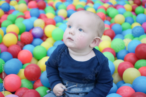 A cute eared baby sits among bright multi-colored balls and looks mysteriously to the side. Cute baby in the children's room. Close-up portrait