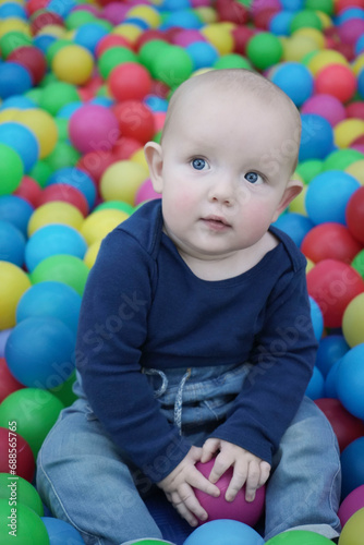 A cute eared baby sits among bright multi-colored balls and mysteriously looks up to the side. Cute baby in the children's room. Portrait. Vertical photo.