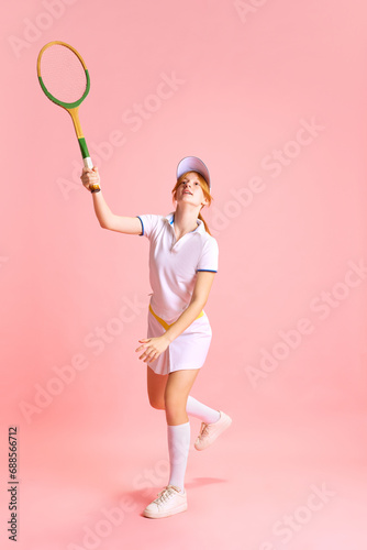 Happy cheerful young lady, dressed retro tennis uniform with racquet getting high while workouting in motion against pastel pink background © Lustre