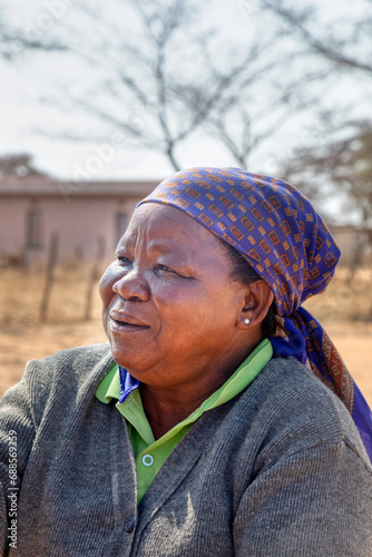 villager , headshot of old african woman with blue scarf, house and dirt road in the background