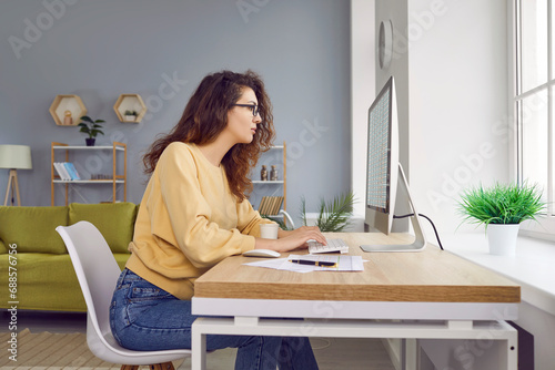 Beautiful young woman accountant is working from home, sitting at her desk, and working with financial spreadsheets on a modern desktop computer. Side view. Remote work concept photo