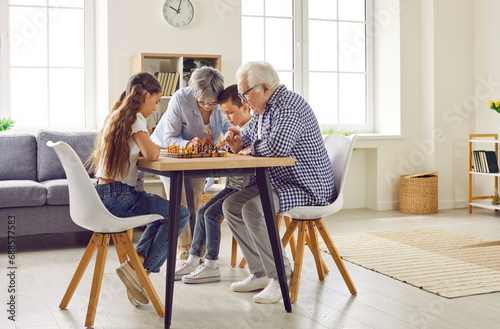 Grandparents teach their grandchildren how to play chess while spending time together at home. Concentrated brother and sister and their grandparents sit in living room at table with chess board.