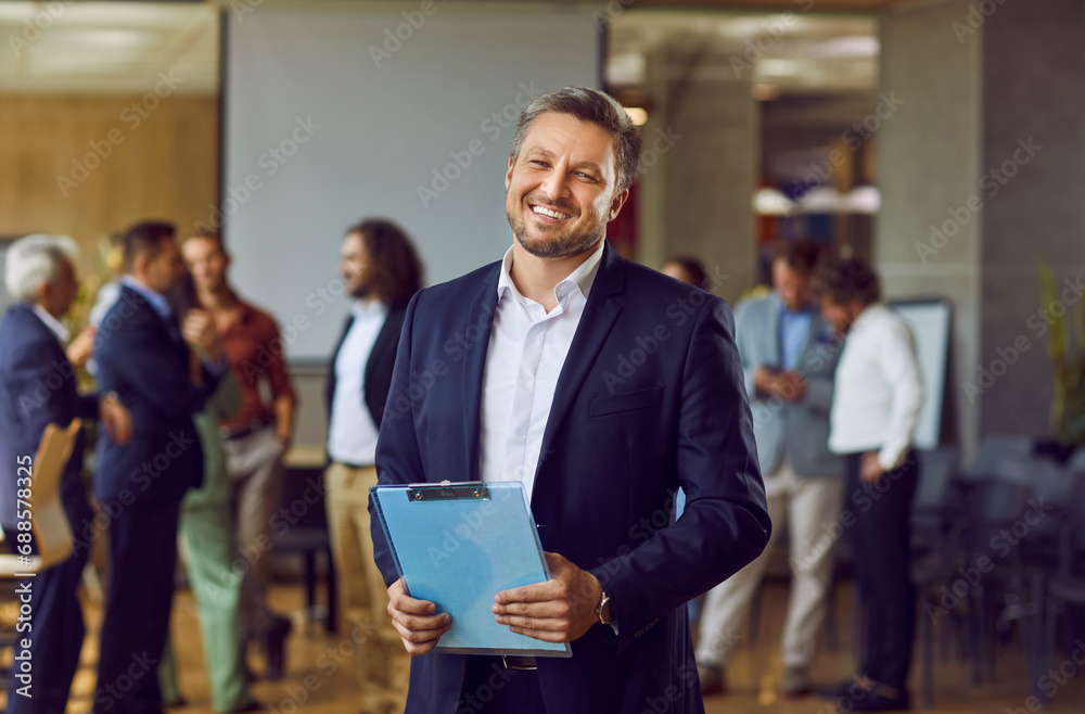 Portrait of happy smiling confident business man looking at the camera holding a folder with financial documents with a team of company employees talking in background in modern office. - obrazy, fototapety, plakaty 