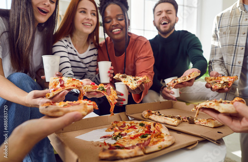 Happy young diverse multiethnic people friends or colleagues having party gathering at home eating slices of pizza from box, talking and laughing together. Delivery service concept.
