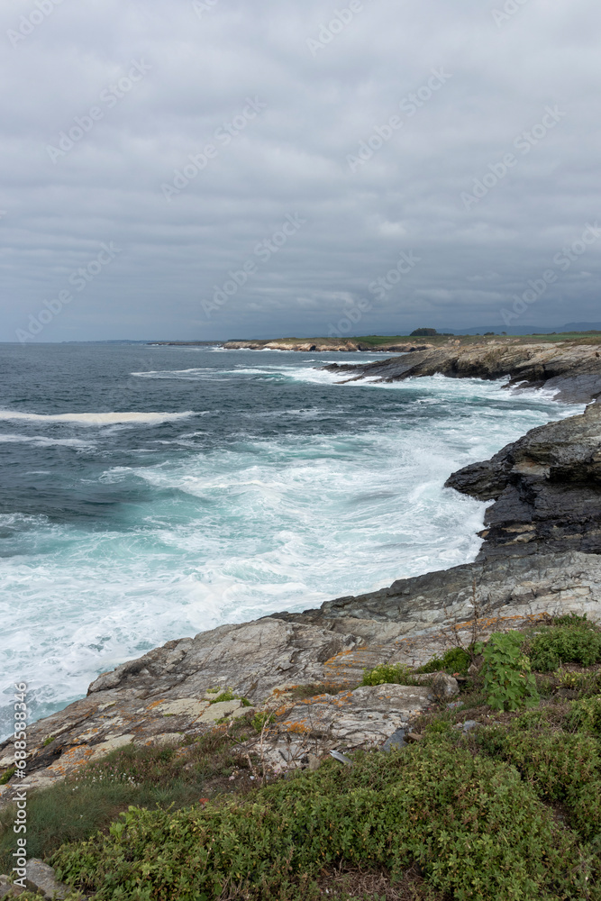A turbulent ocean crashes against a rocky coastline under a cloudy gray sky, with a small island visible in the distance