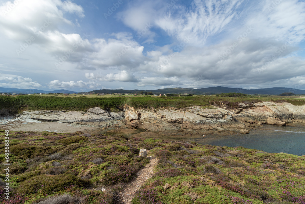 A rocky coastline covered in green and purple vegetation under a bright blue sky with white clouds, with deep blue water visible