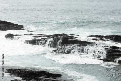 a rocky coastline with light blue, rough waves crashing against black, jagged rocks under an overcast light gray sky