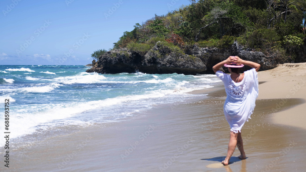 An elegant tourist in a white summer dress and a pink hat walks along the beach on a sunny day on the ocean. He looks into the camera and at the ocean, resting on the shore of a tropical beach.
