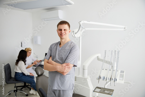 A young male dentist stands in the middle of the office  and in the background a colleague is advising patients.