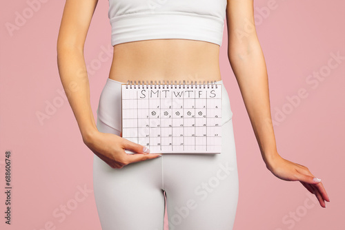 Young caucasian slim lady in sportswear showing calendar on pink studio background, cropped, close up photo