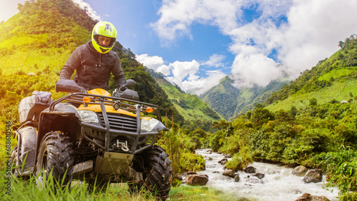 Motorcyclist on quad bike. Man rides ATV off-road. Extreme racing in mountains. ATV racer under summer sky. Quad cyclist in yellow helmet. Driver of quad bike crosses river. ATV near stormy river. photo