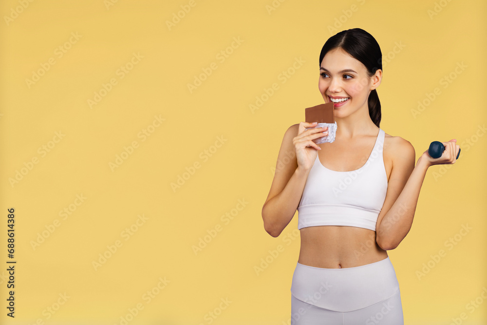 Cheerful caucasian woman in sportswear with dumbbell eating chocolate isolated on yellow studio background Cheerful caucasian woman in sportswear with dumbbell eating chocolate isolated on yellow studio background