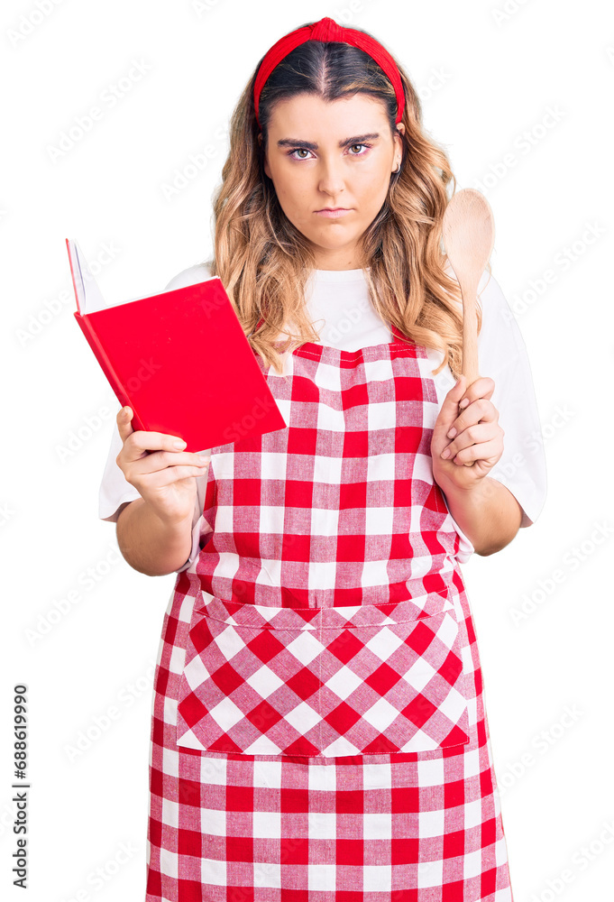 Young caucasian woman wearing apron holding recipe book and spoon thinking attitude and sober expression looking self confident