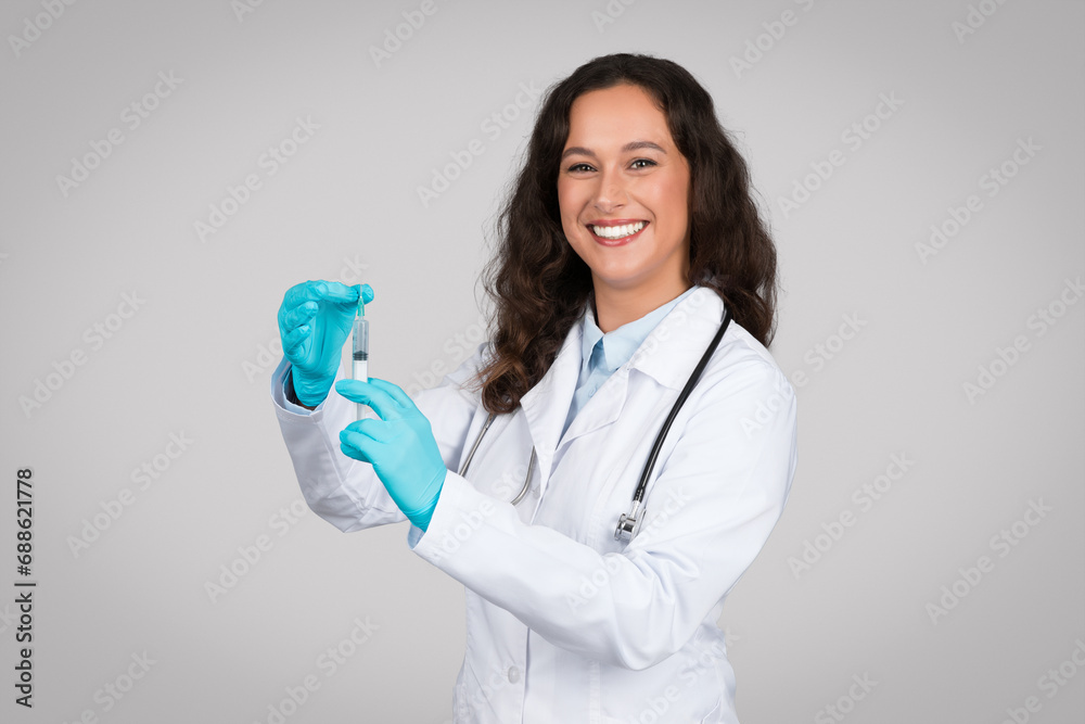 Cheerful lady doctor in white coat, protective gloves holding syringe and smiling at camera on grey studio background Cheerful lady doctor in white coat, protective gloves holding syringe and smiling at camera on grey studio background