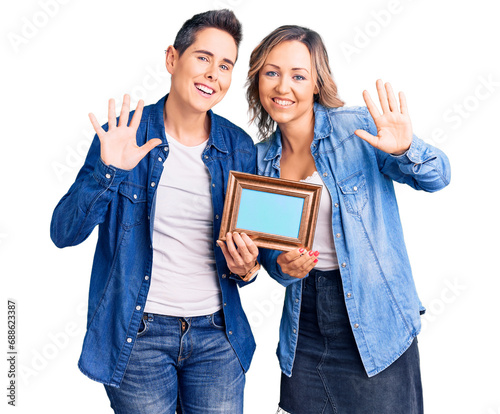 Couple of women holding empty frame waiving saying hello happy and smiling, friendly welcome gesture