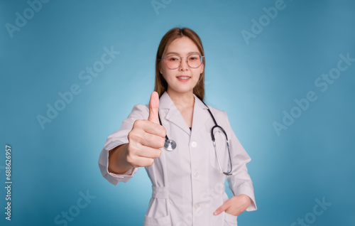 Young asian doctor put hand in pocket of medical uniform lab coat and stethoscope on blue background.. Smiling beautiful female doctor with thumb up. Looking at camera. Hand focus.